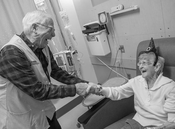 Penn State Health volunteer Bill Moore laughs as he holds the hand of an older woman receiving a chemotherapy infusion at Penn State Cancer Institute. The woman is sitting in a chair and wearing a small witch hat for Halloween. A tube is attached to a vein in her hand. Bill has white hair and is wearing a plaid shirt and a vest with the Penn State Health Volunteer Services logo on it. Behind them is an IV stand with several bags of medicine and a plastic bin on the wall.
