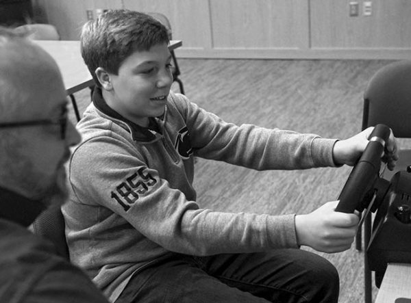 A teen boy smiles and holds the wheel of the driving simulator One Simple Decision. Next to him is a man with glasses, gray hair and a beard. Behind them are a table and chairs, carpet and a wooden cabinet.
