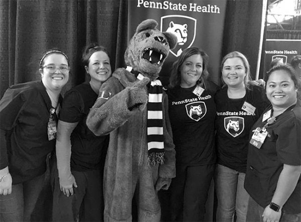 The Penn State Nittany Lion poses with his thumb up and a bandage on his arm at the Pennsylvania Farm Show. He is standing with five women who are community health nurses at Penn State Health Milton S. Hershey Medical Center. Behind them is a Penn State Health banner and a curtain.