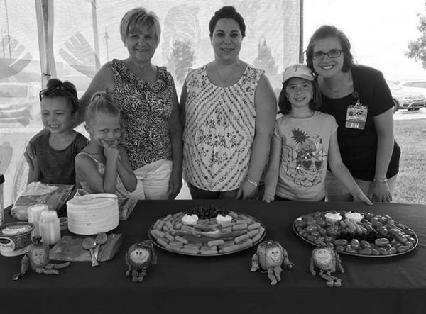 Three women smile at a table with three young girls at the Farmers Market in Hershey in 2017. Two trays of vegetables are on the table in front of them with four dolls shaped as fruits and vegetables. Behind them is a thin curtain. A row of parked cars is on the right.