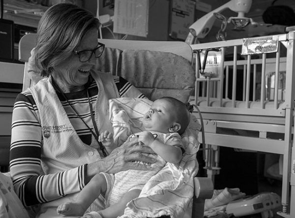Lori Brullo, a volunteer with patient services at Penn State Health Milton S. Hershey Medical Center, smiles and holds a baby at Penn State Children’s Hospital. She is sitting in a padded rocking chair and wearing a striped shirt and vest with the Penn State Hershey Volunteer Services logo on it. She has short hair and glasses. The baby is wearing a onesie and is lying on a blanket and looking up. Behind them is a crib with a mobile in it.