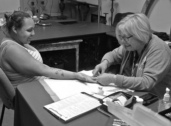 A woman community health nurse from Hershey Medical Center does a finger stick test on a woman at a food pantry. The nurse is wearing glasses, a sweater, scrubs, a Penn State Health lanyard and rubber gloves. The client is smiling as she stretches out her arm on a paper towel. She is wearing a striped tank top and has tattoos on her back and arm. The table has a clipboard, phone and a plastic container on it. Behind them are tables with various household items on them.