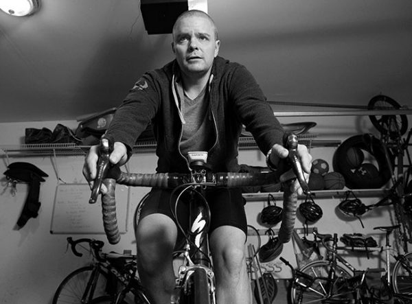 Tim Card of Mount Joy, Pa., the first patient at Penn State Cancer Institute to receive CAR-T therapy, rides on a stationary bike in his garage. He is wearing a sweatshirt, T-shirt and bike shorts. Behind him is a shelf filled with sports equipment and bike helmets on the wall. A garage door opener and a light are above him.