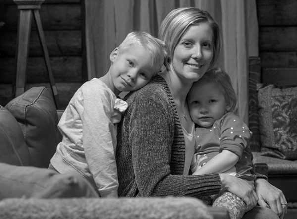 Krista Carroll of Shippensburg smiles with her children Madden, 6, and Jillian, 3, in a black and white portrait. They are sitting on a couch. Krista is wearing a sweater and casual top. Madden is leaning his head on his mother’s back and is wearing a long-sleeved shirt. Jillian is sitting in her mother’s lap and wearing a polka-dotted shirt. Behind them is a log cabin wall, a curtain and pillows on a chair.