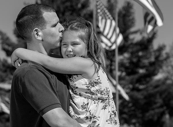 Paul Brennan kisses his daughter Lilly’s forehead as he holds her in his arms. They are standing in front of a row of American flags. He has short hair and is wearing a polo shirt. She has shoulder-length hair, is wearing a flowered dress and is smiling. Behind them are trees.