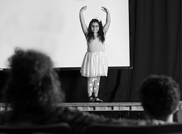 Gabriella Asadi dances on a stage, smiling at her mother and brother. The backs of their heads are shown from behind as they sit in their audience seats.