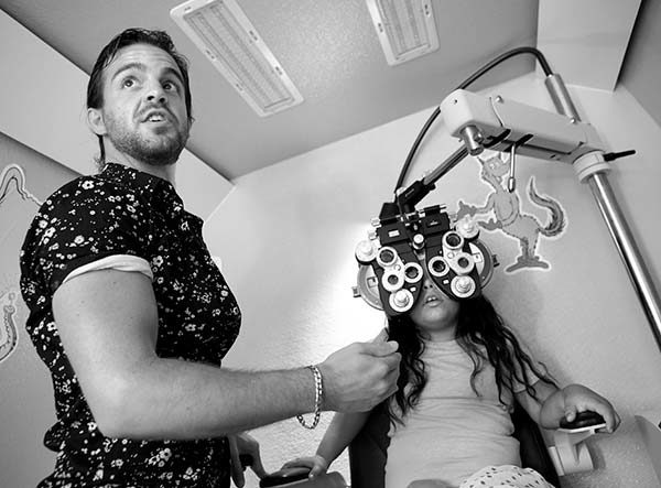 Dr. Aaron Harsch, an optometrist with Vision to Learn, tests Gabriella’s vision with a piece of equipment with many lenses positioned in front of Gabriella’s face as she sit in a chair in the mobile lab. Harsch wears a flowered lab coat and has a beard.
