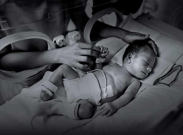 A premature infant lies sleeping in an isolette inside a neonatal intensive care unit. The infant has dark hair and is wearing a diaper, socks and an identification bracelet. A monitor is attached to the infant’s abdomen. A woman’s hands are extended through the arm holes and touching the infant’s head and hand.