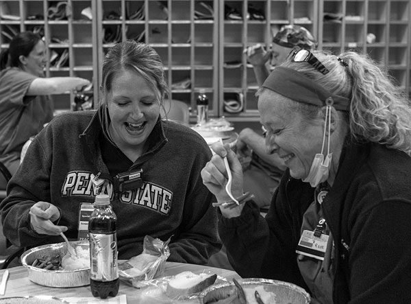 Jennifer Gramley, nurse practitioner, and Kate Perkins, registered nurse, sit at a round table, eating ham, potatoes and string beans from foil food containers. Gramley wears a Penn State pullover and has her hair in a ponytail. Perkins has a ponytail and wears a jacket over scrubs and a face mask under her chin while she eats.