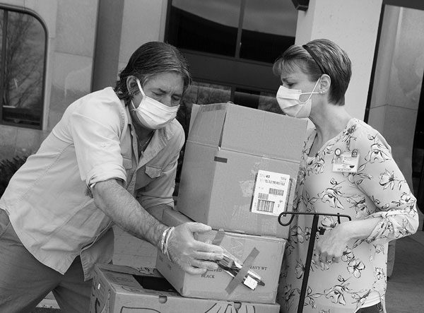 Phil Guano loads four boxes of Easter meals onto a wheelchair in a parking lot. Tracy Johnsen has her hands on the wheelchair’s handles. Both are wearing masks. The hospital entrance is in the background.