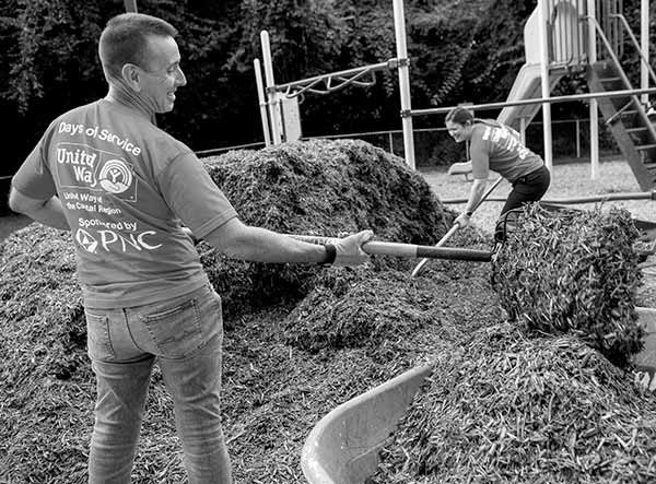 Dr. Kevin Black, interim dean of the College of Medicine, and Deborah Berini, president of Penn State Health Milton S. Hershey Medical Center use pitchforks to scoop mulch outdoors at the YWCA playground. They are wearing United Way T-shirts and jeans. Behind Deborah is a jungle gym.