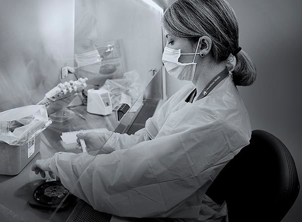 A woman medical laboratory scientist at Penn State Health Milton S. Hershey Medical Center holds a test tube as she performs a test. On the counter in front of her is a container lined in plastic and lab equipment. She is wearing plastic scrubs, a face mask and a lanyard around her neck. She has short hair pulled into a ponytail.