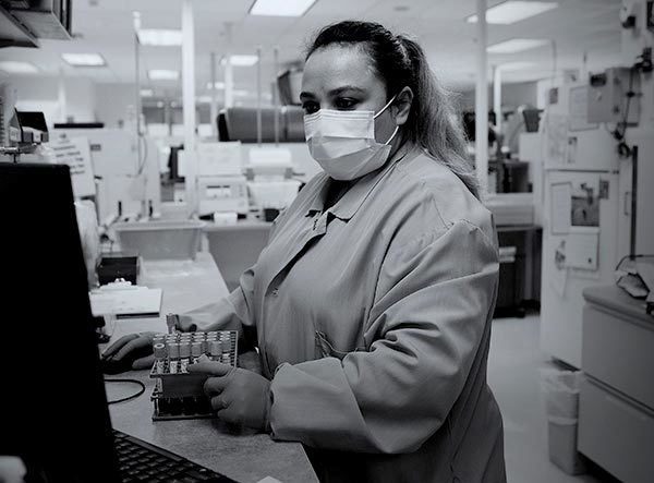 A woman medical laboratory scientist at Penn State Health St. Joseph touches a box of test tubes as she looks at a computer screen. She is wearing scrubs and a face mask and has long hair in a ponytail. Behind her are lab equipment and lights.