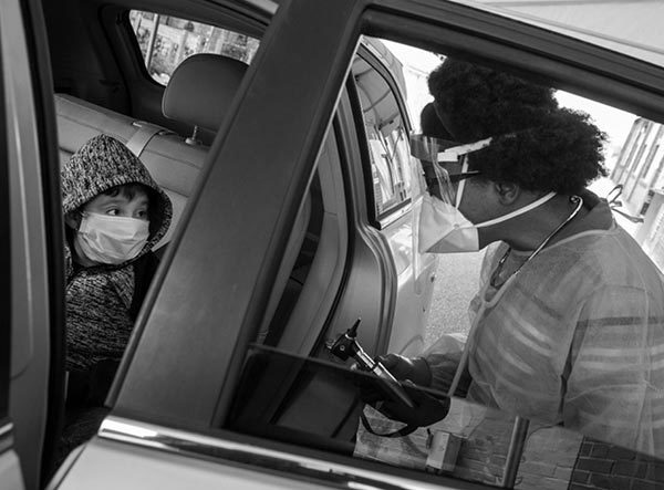 Dr. Desiree Webb, wearing a gown, face mask and stethoscope, stands next to a car with a boy who is wearing a face mask and jacket. The car door is open.