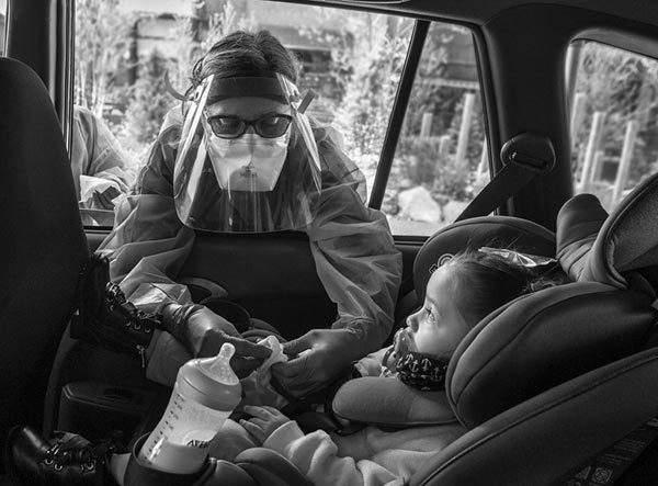 Penny Porter a nurse practitioner with Penn State Health Medical Group ― All About Children, leans into a car and touches the fingers of 2-year-old Zyann Perez-Cruz. Porter is wearing a gown and face shield. The child is in a car seat and has a bottle in the cup holder next to her.