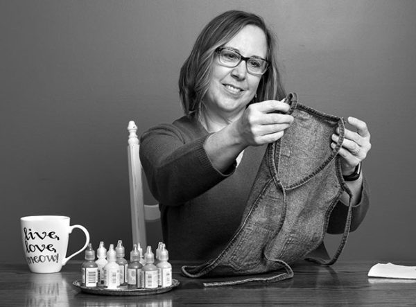 Wendy Johnson, who has shoulder-length hair and wears glasses, sits behind a table and smiles as she looks at a child’s apron she holds in front of her. Bottles of fabric paint and a mug that says “Live, Love, Meow” are on the table.