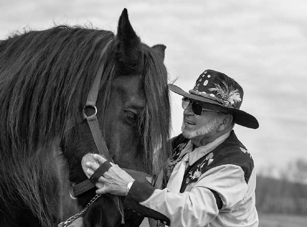 A man smiles as he holds the harness of a horse. The man has a beard and is wearing sunglasses, a cowboy hat with a feather in it, a flowered vest and a long-sleeved shirt. The horse’s head is visible with a long forelock hanging over its eyes and a harness on the right side of its head.