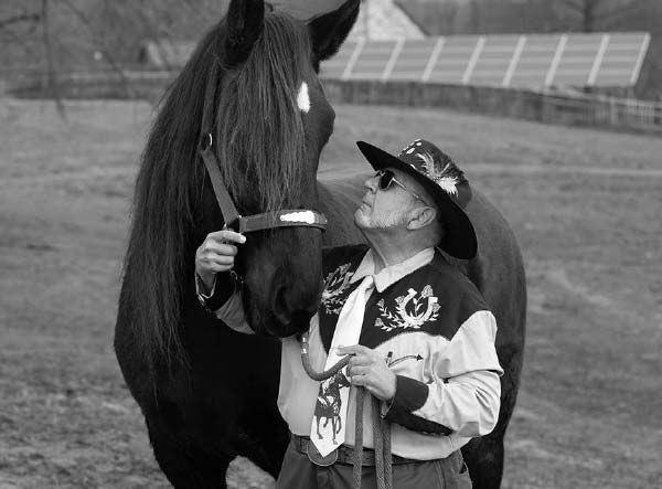 A man looks up at a horse and holds its harness in his right hand and a rope in his left hand. The man has a beard and is wearing sunglasses, a cowboy hat with a feather in it, a flowered vest and a long-sleeved shirt. The horse’s entire body is visible. It has a spot on its forehead, and its forelock hangs over its eyes. Behind them a fenced arena and solar panels are visible.