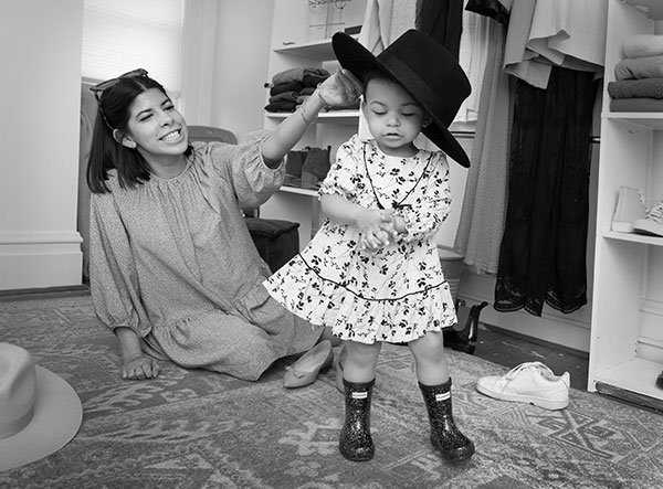 Alyssa Acevedo, who has shoulder-length hair and wears a long-sleeve dress, sits on her bedroom floor and smiles as she put a large felt hat on her toddler daughter’s head. Izzy, who wears a print dress and boots, clasps her hands together. Shoes and clothes surround them on closet shelves and on the floor.