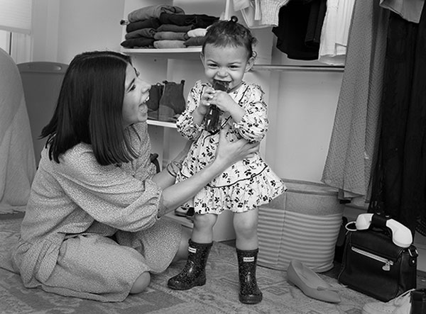 Alyssa Acevedo, who has shoulder-length hair and wears a long-sleeve dress, hugs her toddler daughter, who stands in front of her. Izzy wears a print dress and chews on sunglasses. Shoes and clothes surround them on closet shelves and on the floor.