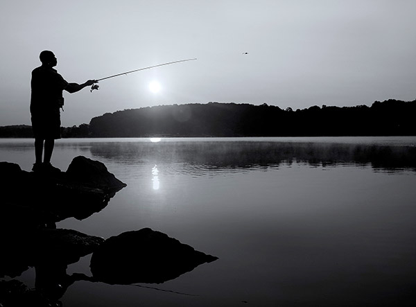 Silhouette of a man facing right, standing on a rock by the water’s edge, casting a fishing line out into a lake. The sun is setting behind a hill covered in trees and is reflected in the lake.