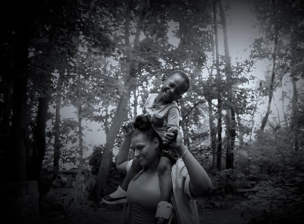 Mother smiles as son sits on shoulders and laughs while holding mother’s hands in a wooded setting.
