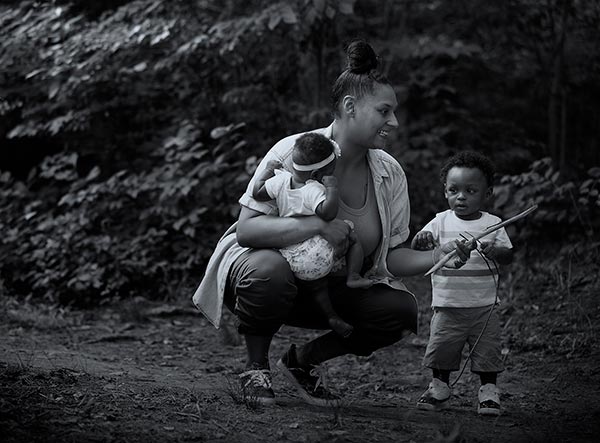 Mother squats to talk to small child while holding baby in her right arm on a wooded trail.