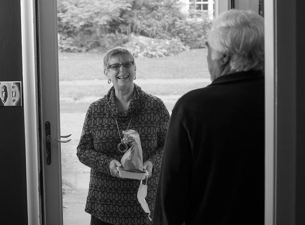 A woman stands in a doorway on a porch holding a bag and a package of food. She smiles at a man in the foreground who is inside the house and facing her from the doorway. Behind her bushes obscure another house across a swath of grass and pathways.